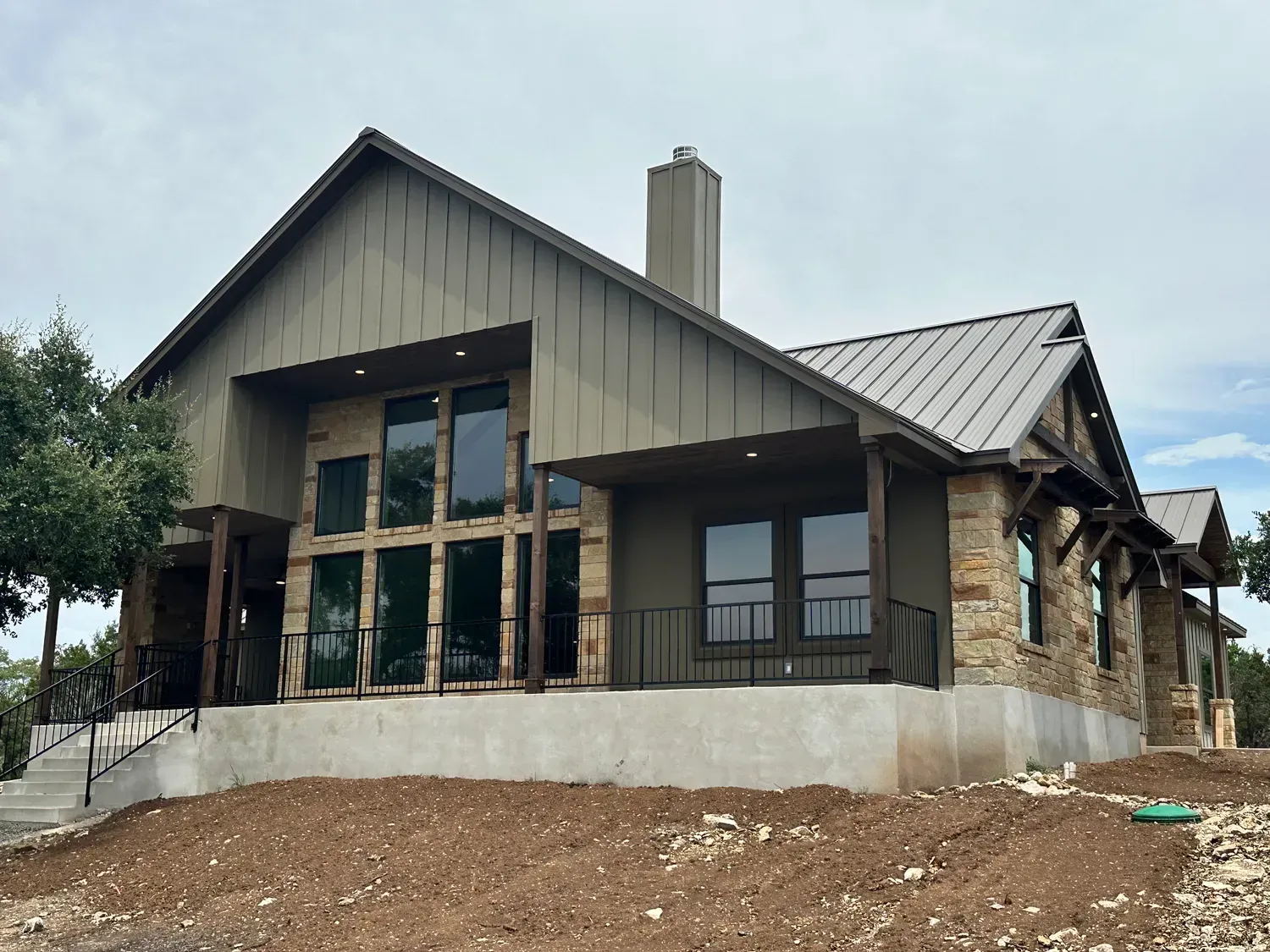 Modern house under construction with gray siding, large windows, and stone accents on a dirt lot