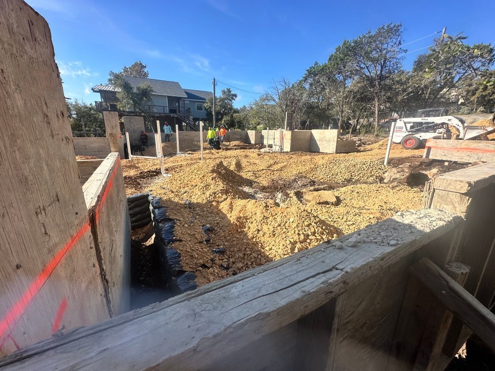 Construction site with excavated dirt pit, wooden framing, and houses in the background under a blue sky