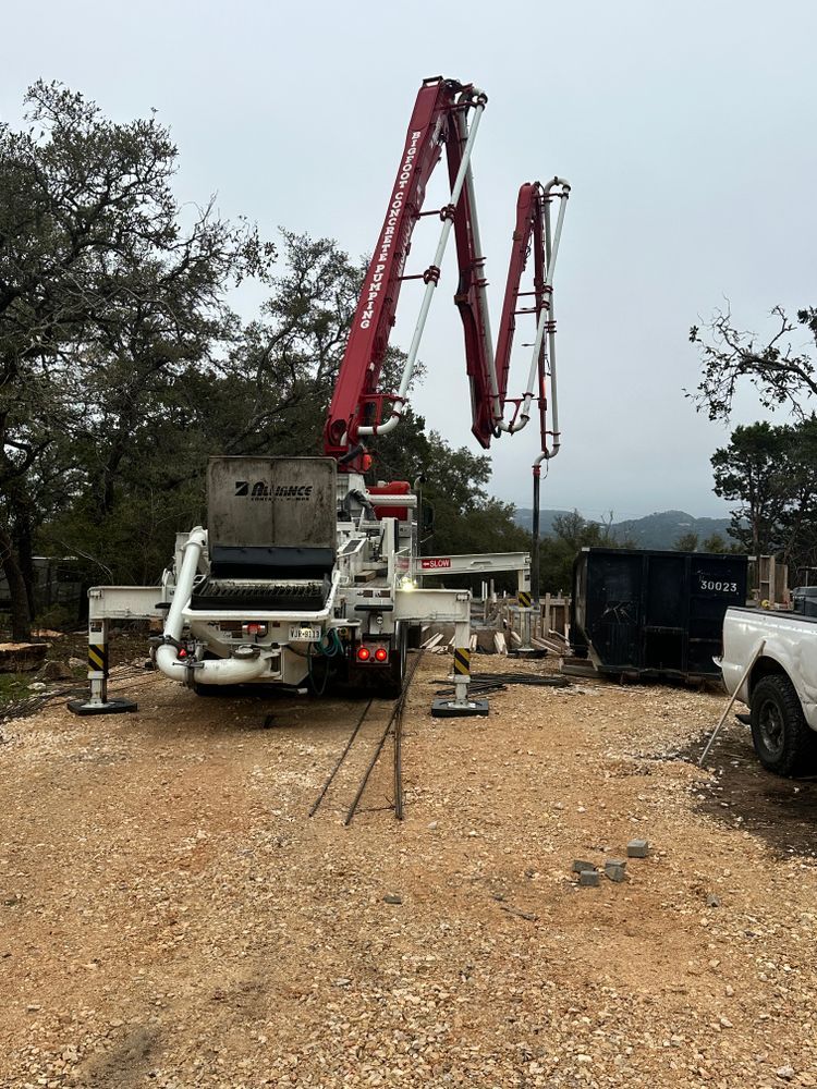 Concrete pump truck with red boom arms at a construction site on gravel, with trees and equipment nearby