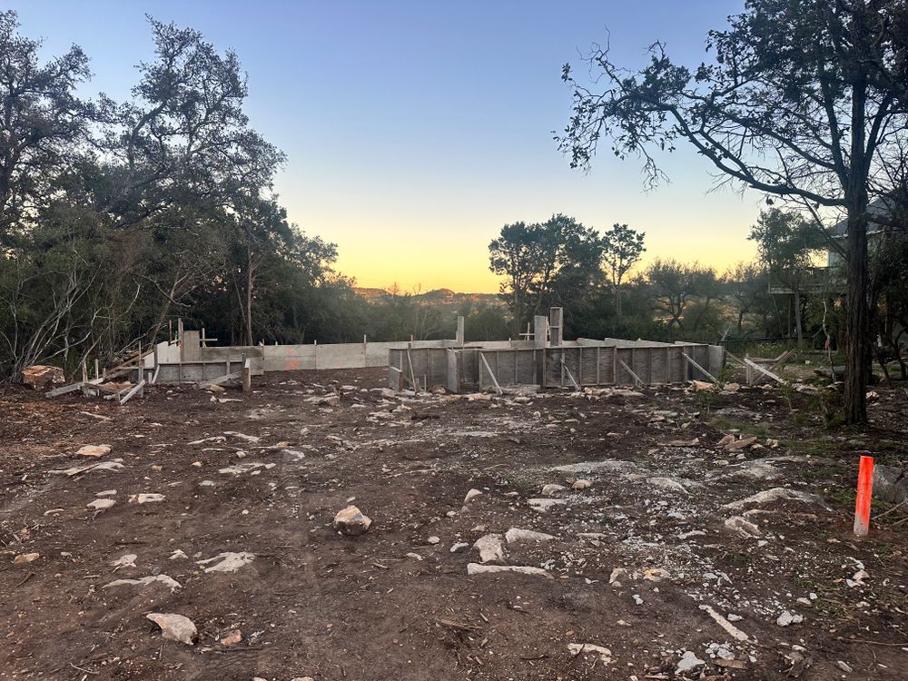 Cleared construction site at dusk with trees, a low wall, and scattered debris under a fading sky