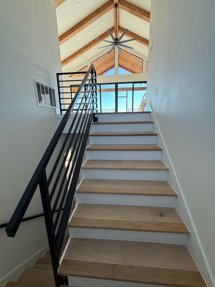 Staircase with black railing leading to a bright loft with exposed wooden beams and large windows