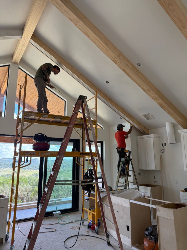 Two workers installing wooden ceiling beams in a bright kitchen with ladders and cabinets.