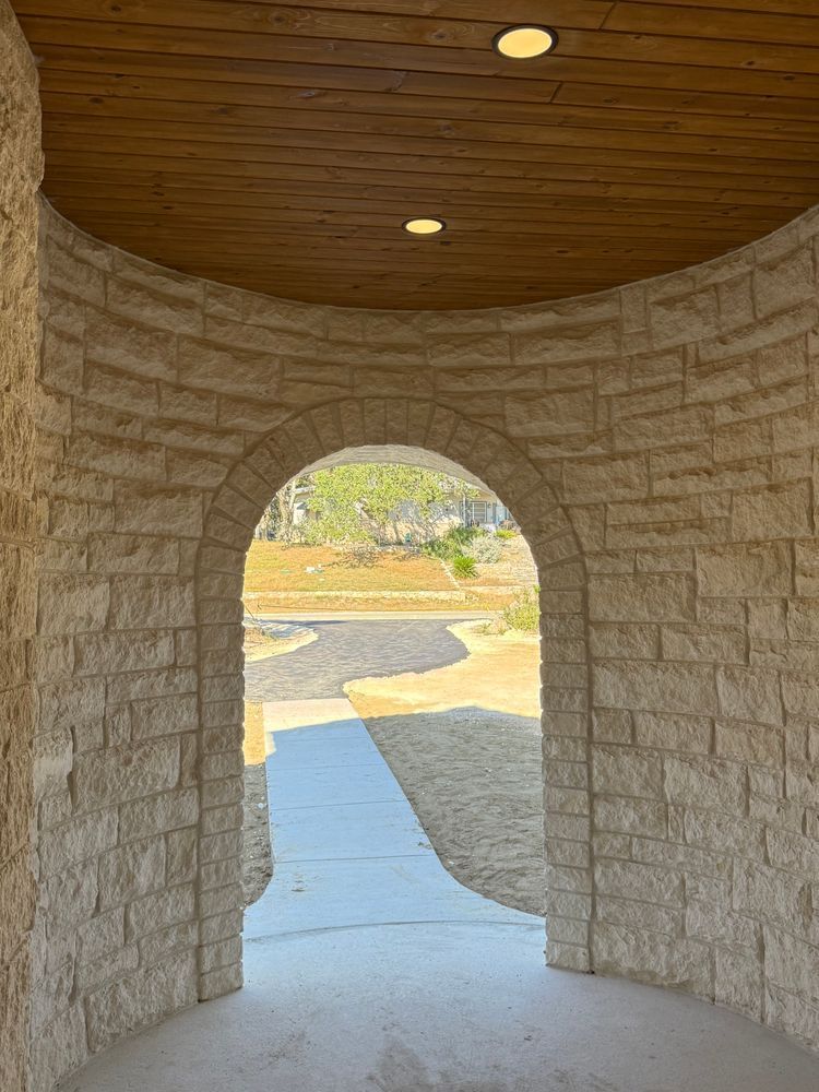 Stone archway opening to a sunlit path and landscaping outside, with a curved wood ceiling overhead