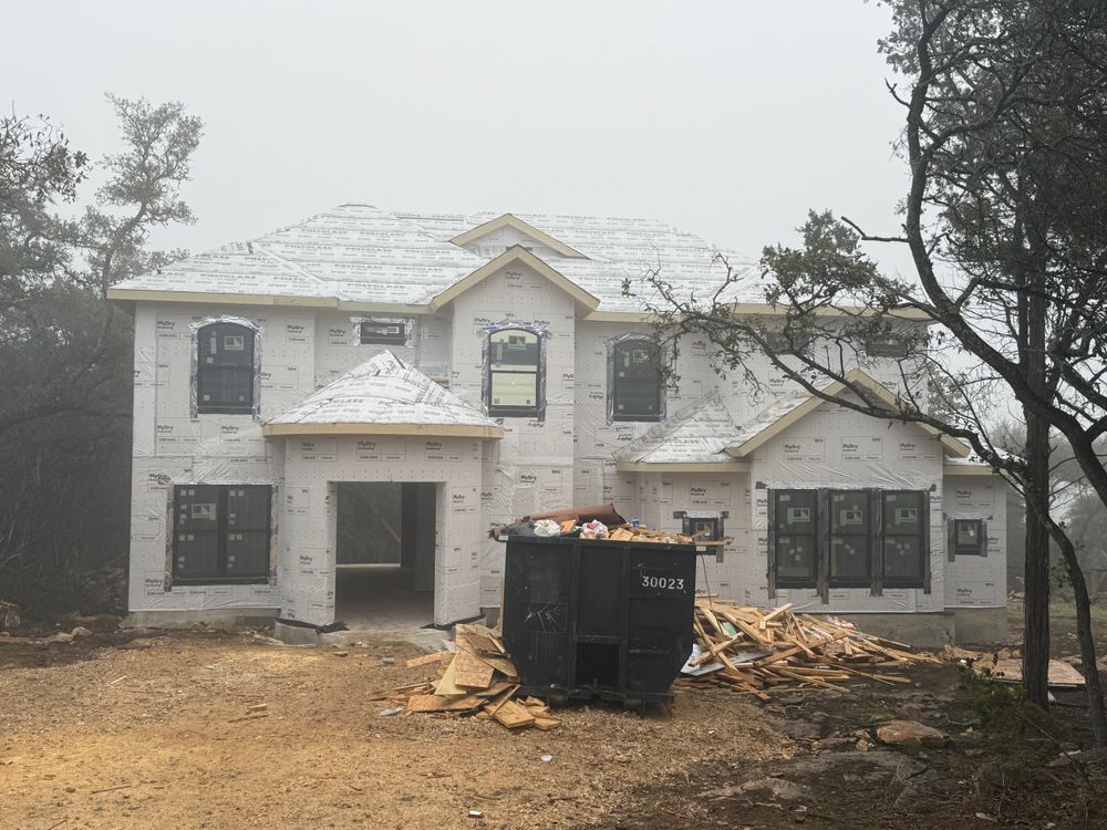 Two-story house under construction with white siding, a dumpster, and lumber piles in front.