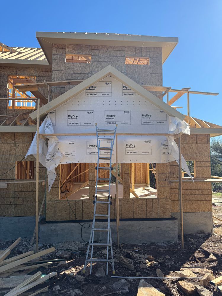 Partially built house with white house wrap, ladder, exposed framing, and dirt foreground