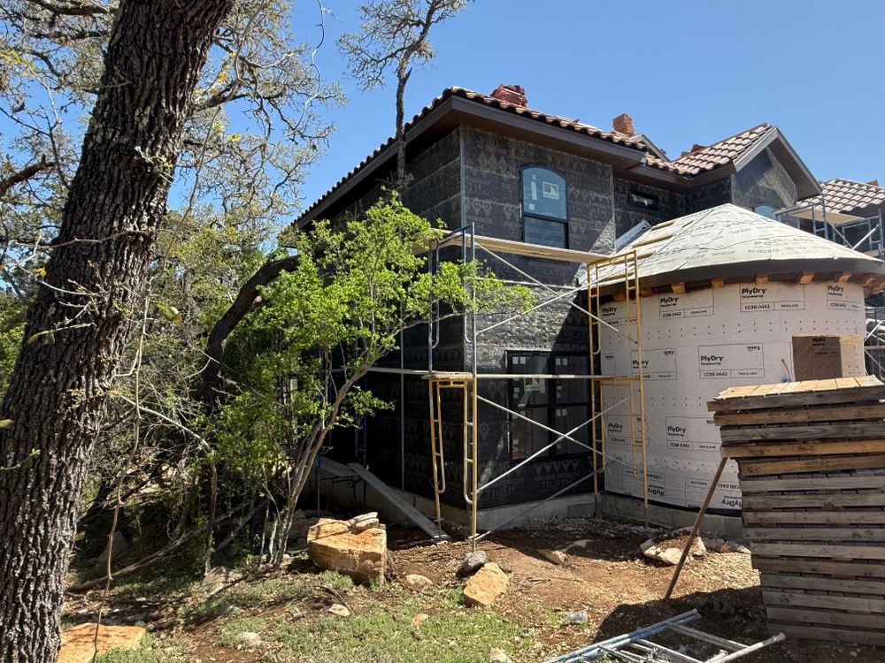 House under renovation with scaffolding, white wrap, and stacked lumber beside a tree in a yard