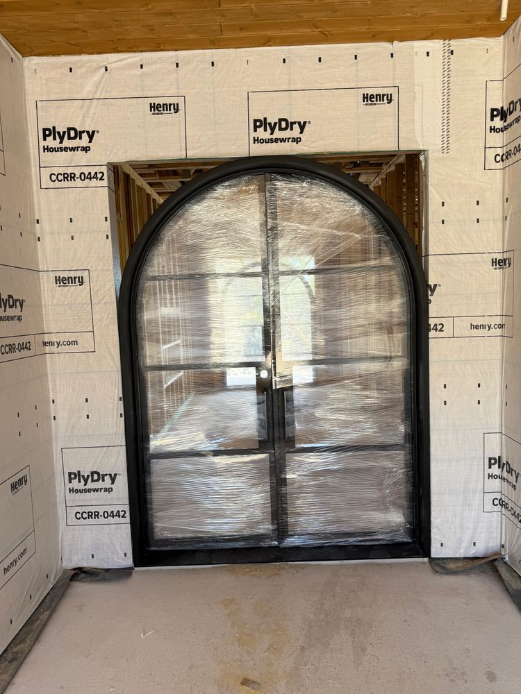 Arched black-framed opening wrapped in plastic in an unfinished room with insulation board walls and concrete floor