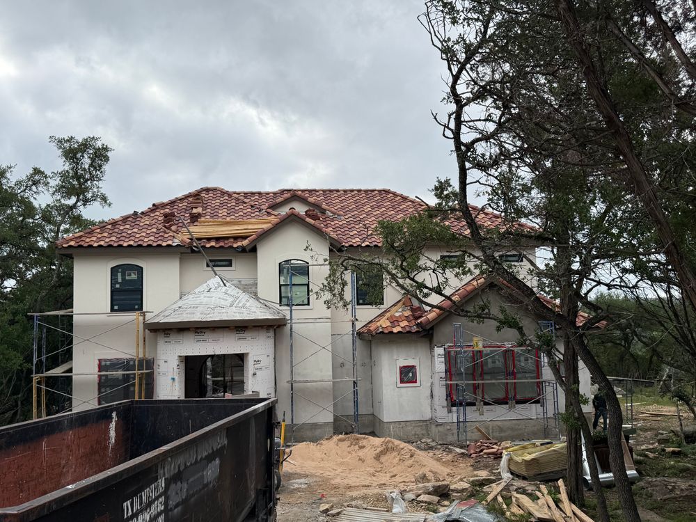 House under construction with red-tile roof, white walls, scaffolding, and sandy ground under cloudy skies
