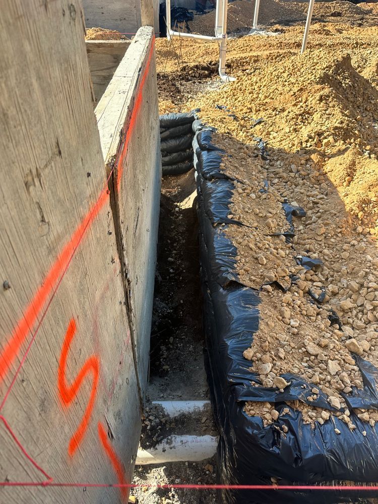 Construction trench beside concrete wall, with black drainage pipe and dirt rubble.