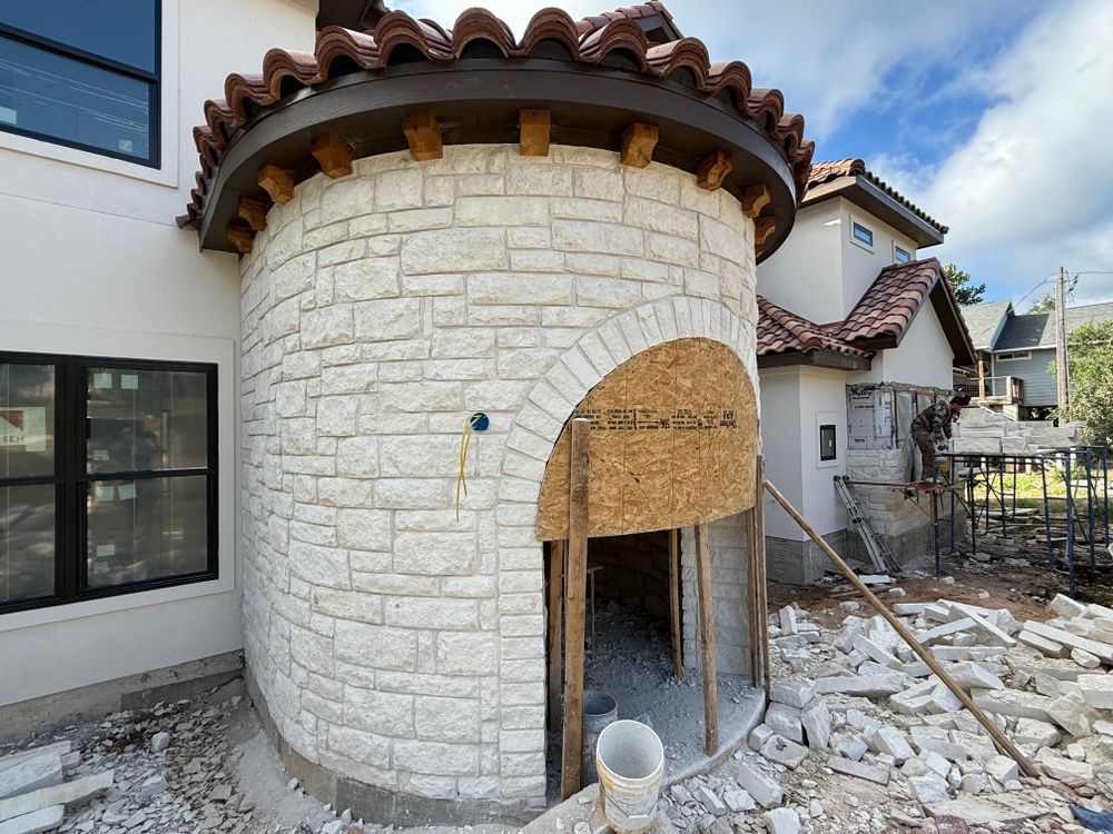 Stone exterior of a house under construction with a rounded turret and open arched doorway, surrounded by rubble