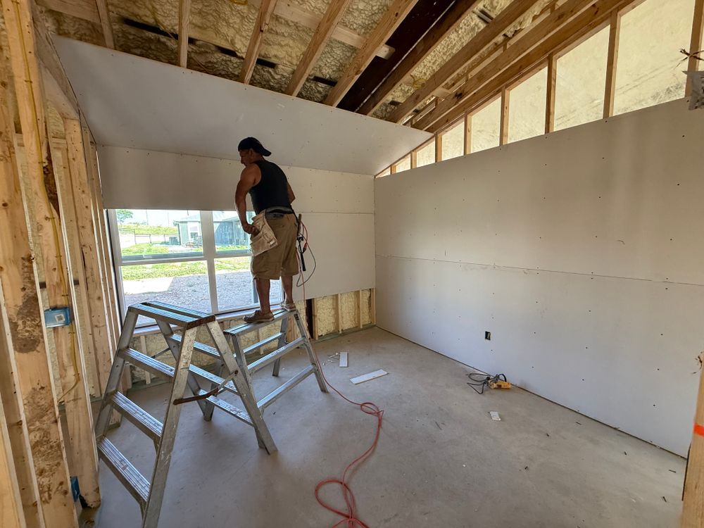 Person on a ladder installing drywall in an unfinished room with exposed framing