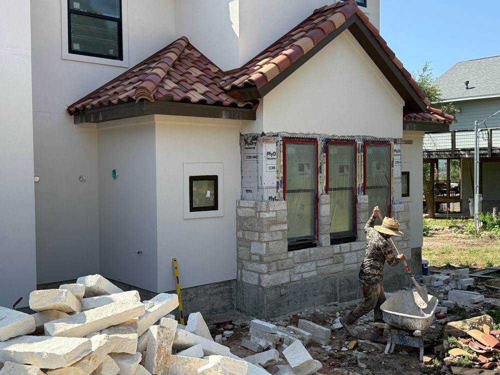 Worker masonry around a house entrance with stone veneer, piles of stone blocks, and construction debris.