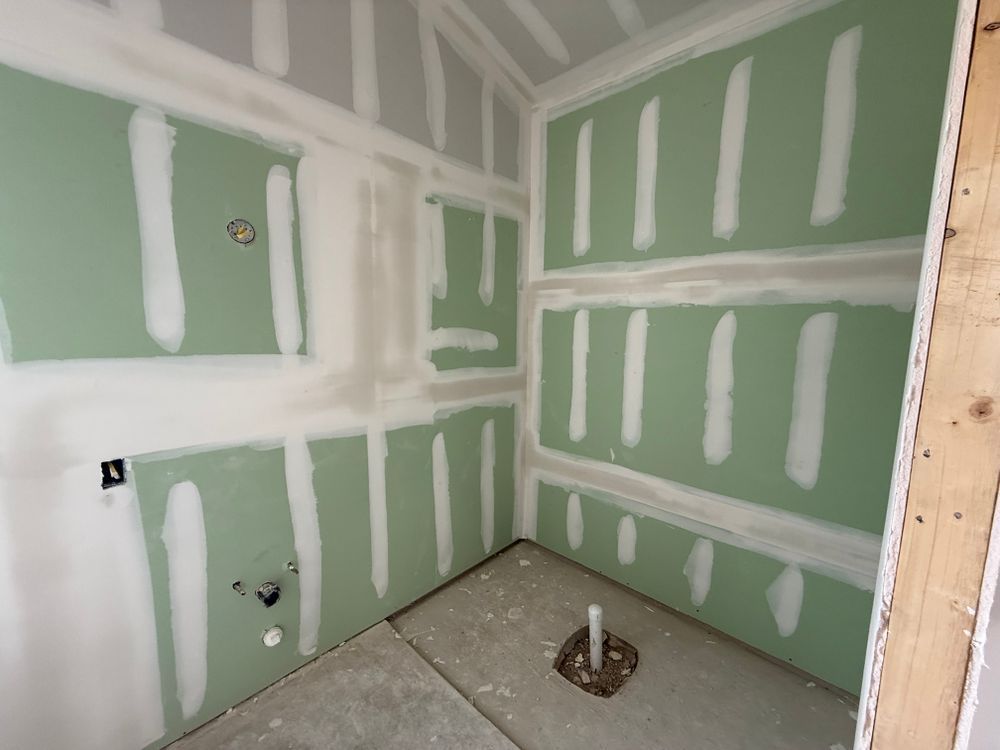 Unfinished room with green drywall, white joint compound, and a bare floor around a plumbing pipe.