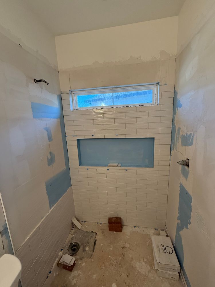 Partially renovated small bathroom with tiled shower wall, blue drywall patches, and a narrow window.