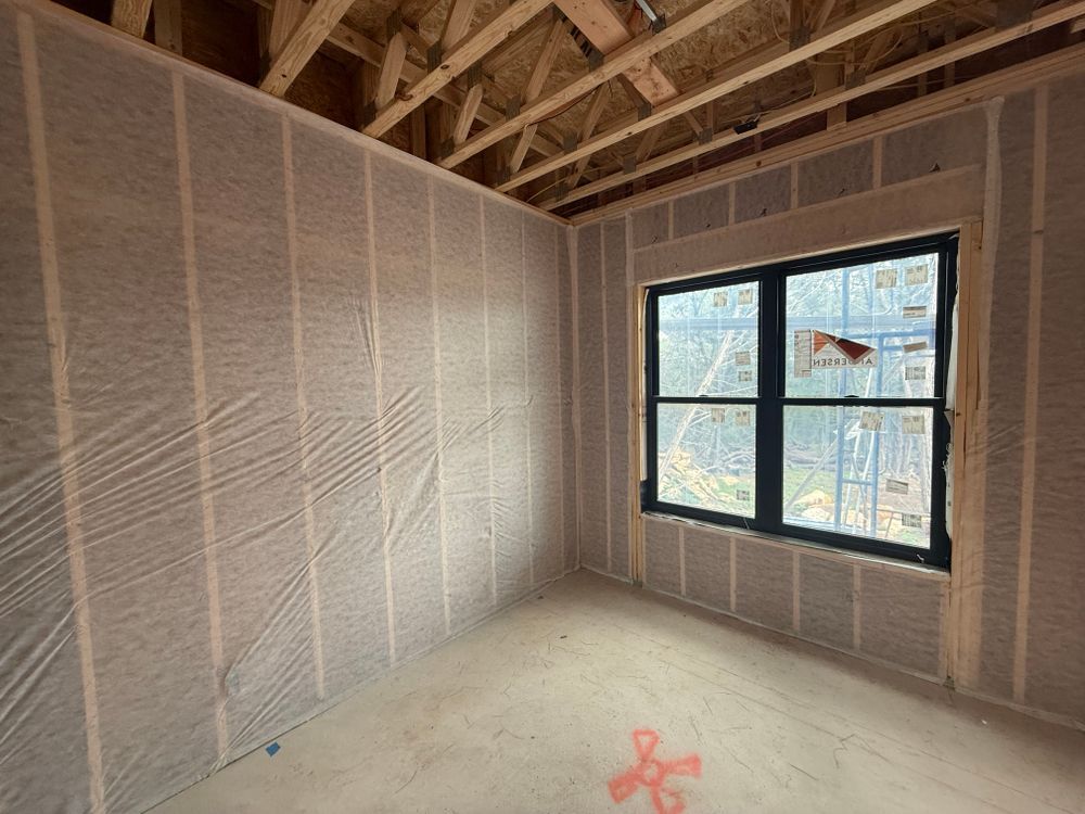 Empty unfinished room with exposed insulation and black-framed window, viewed from a corner.