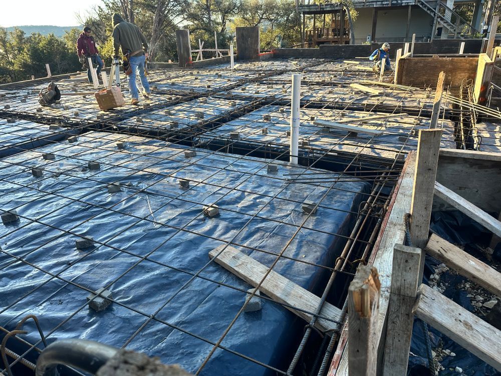 Concrete foundation under construction with rebar grid, black vapor barrier, and workers on site