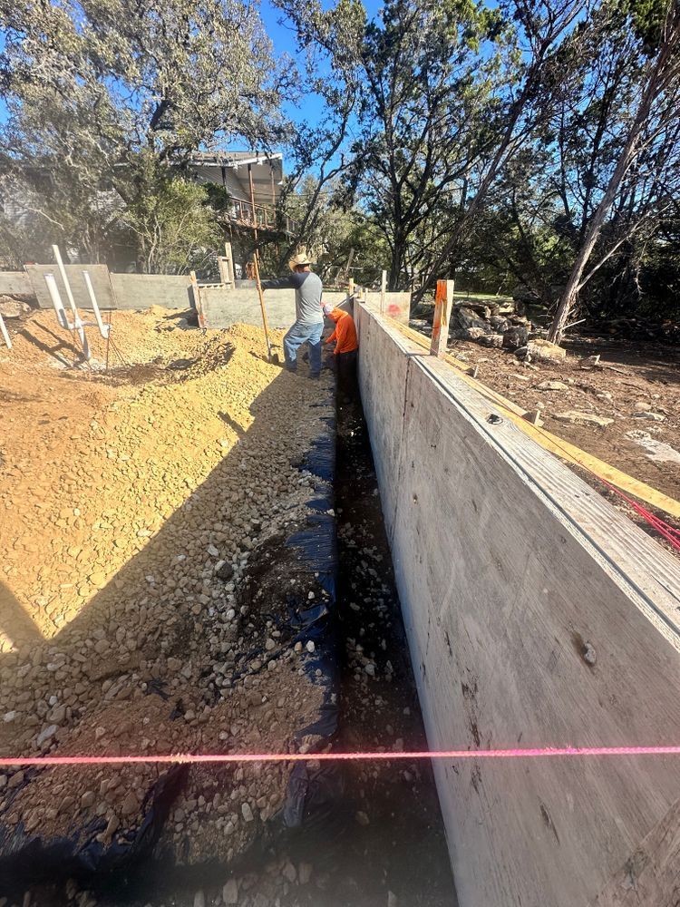 Construction trench beside a concrete retaining wall, with workers and trees in the background.