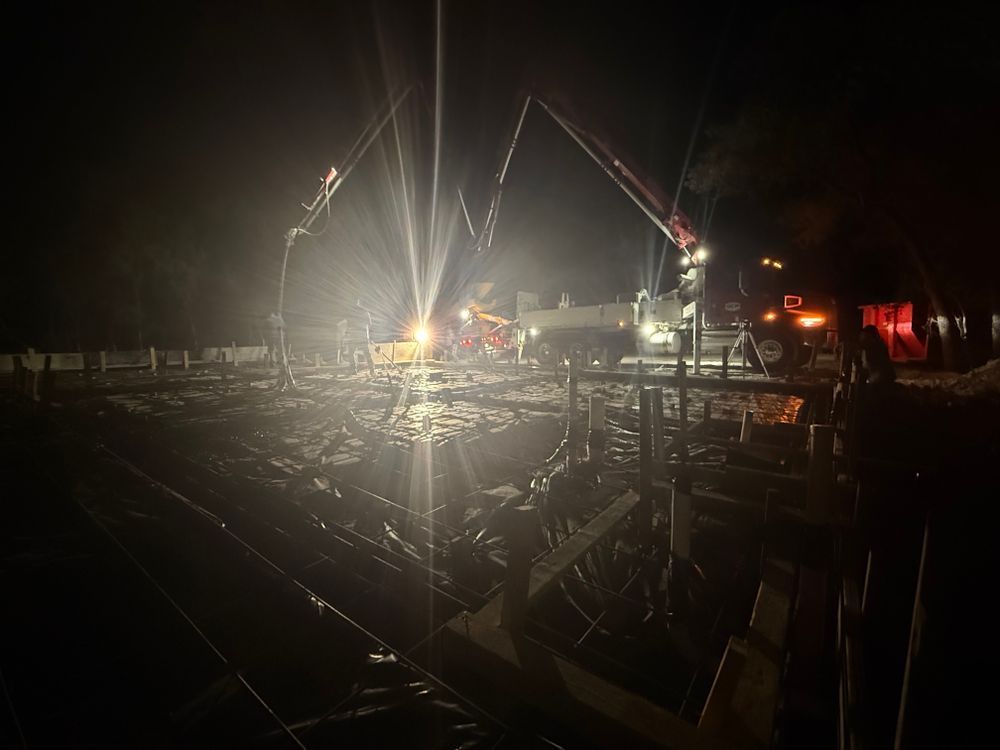 Nighttime waterfront construction site with bright floodlights, cranes, and dock structures over dark water