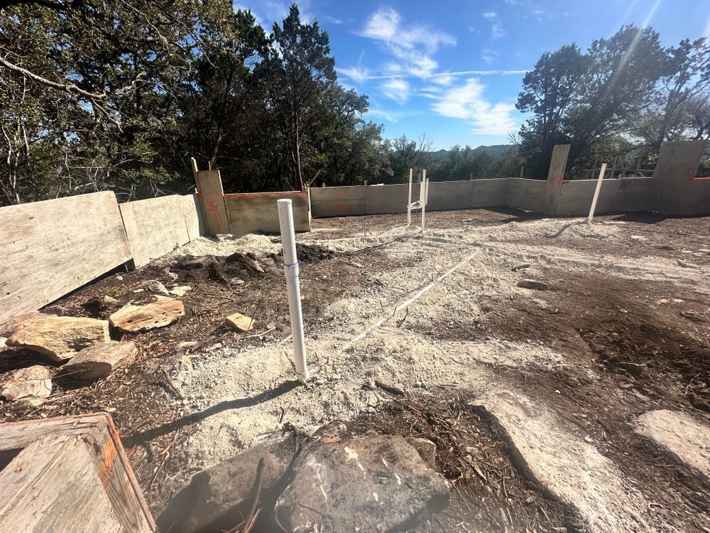 Vacant construction lot with concrete footings, white posts, and trees under a bright blue sky