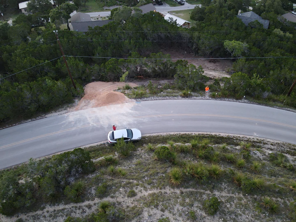 Aerial view of a winding road with a white car stopped and two people nearby in a wooded area.