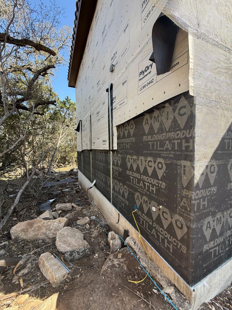 Construction site exterior with house wrap, small window, and rocky ground beside trees