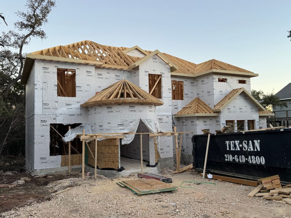 Two-story house under construction with exposed framing, white wrap, and a black dumpster in front