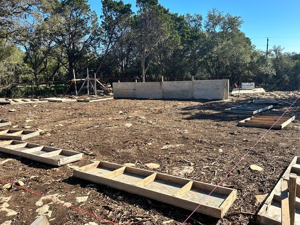 Outdoor construction site with concrete blocks, dirt ground, and a low wall under trees