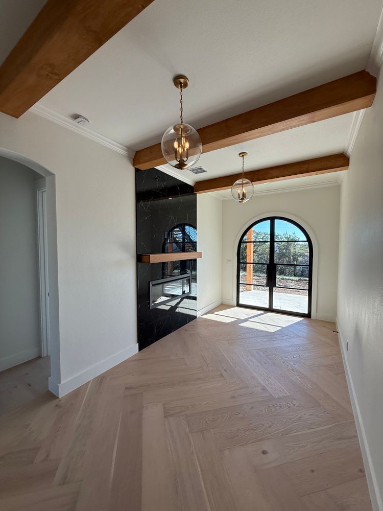 Bright empty room with wood beams, chevron floor, black fireplace wall, and arched glass doors to a patio