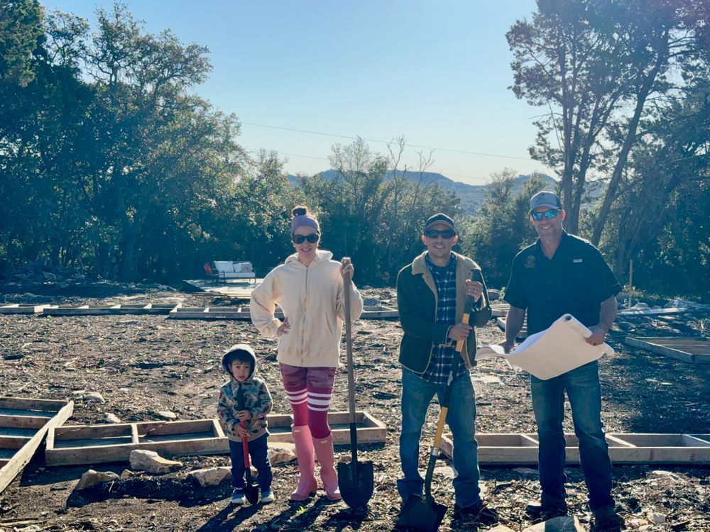 Four people stand on a debris-covered lot, holding papers, with trees and a bright sky behind them.