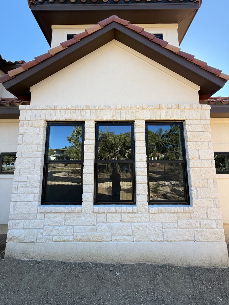 Front exterior of a house with white stone walls, three black windows, and a brown gabled roof.