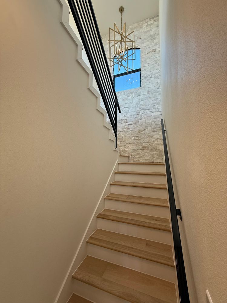 Narrow indoor staircase with light wood steps, black railing, textured wall, and hanging chandelier at top