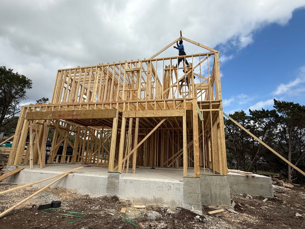 Wood-frame house under construction with worker on roof, cloudy sky, and concrete foundation