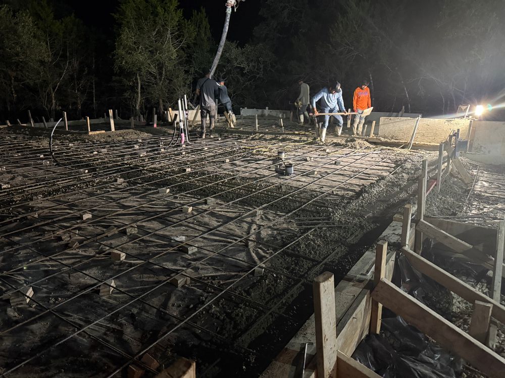 Workers inspecting a rebar foundation at night under bright lights, with concrete forms and soil in the foreground