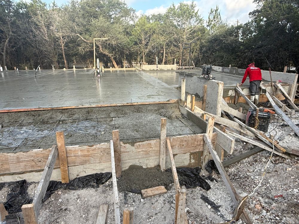 Concrete foundation under construction beside a wet slab, with workers and trees in the background