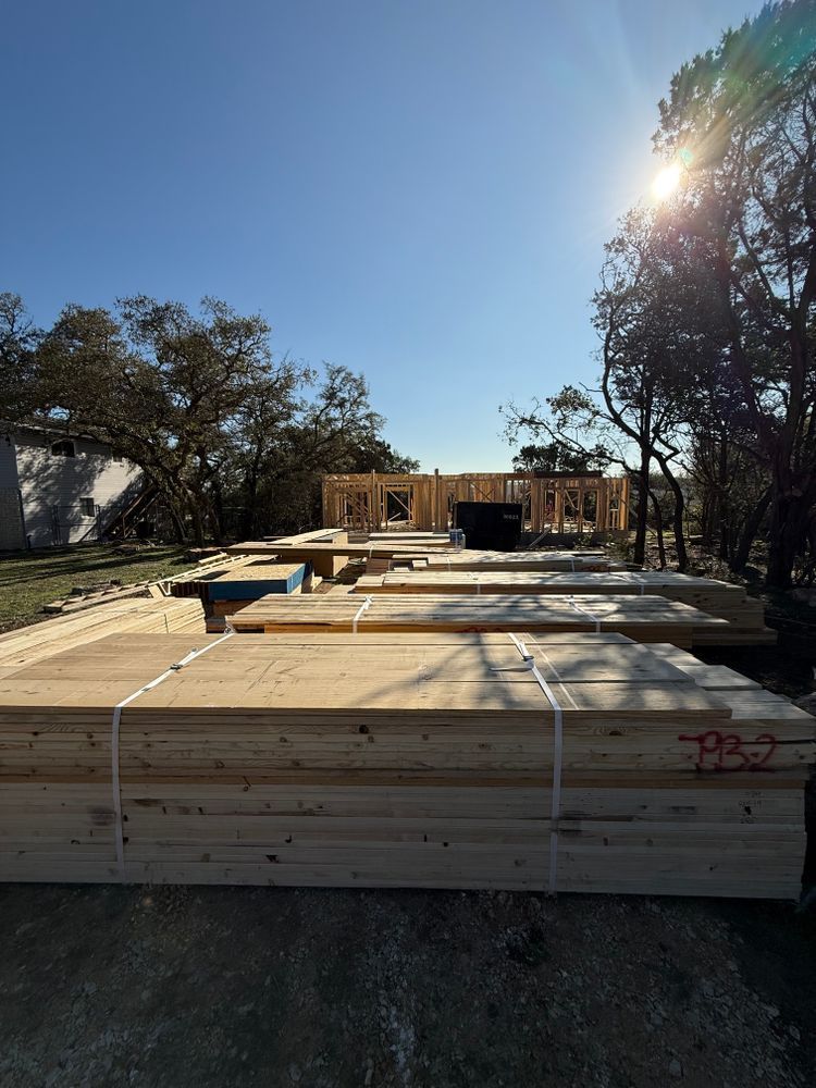 Stacks of lumber at a construction site under a bright blue sky, with trees in the background.