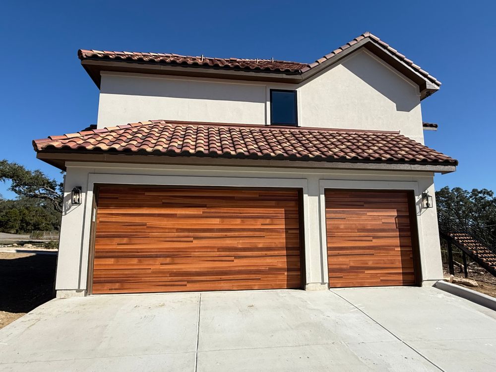 Two-story stucco house with red tile roof and double wooden garage doors in bright sunlight