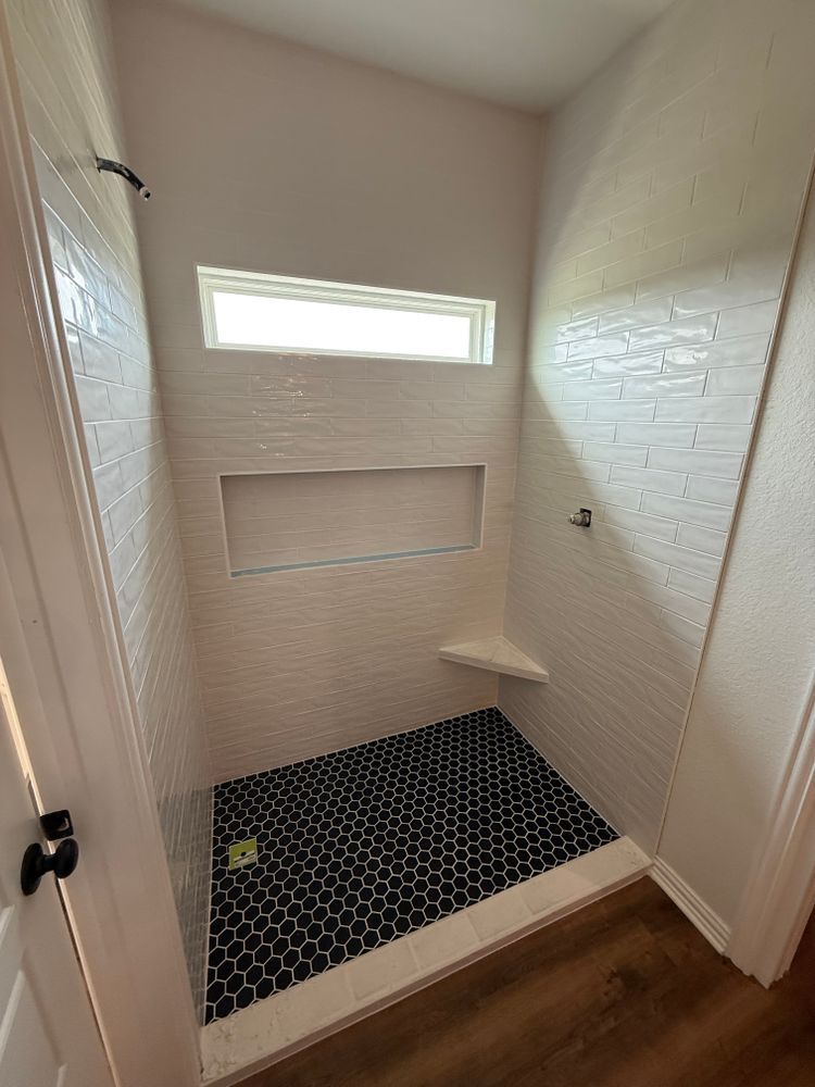 Small tiled walk-in shower with black hex floor, white walls, narrow window, and wood bathroom floor nearby