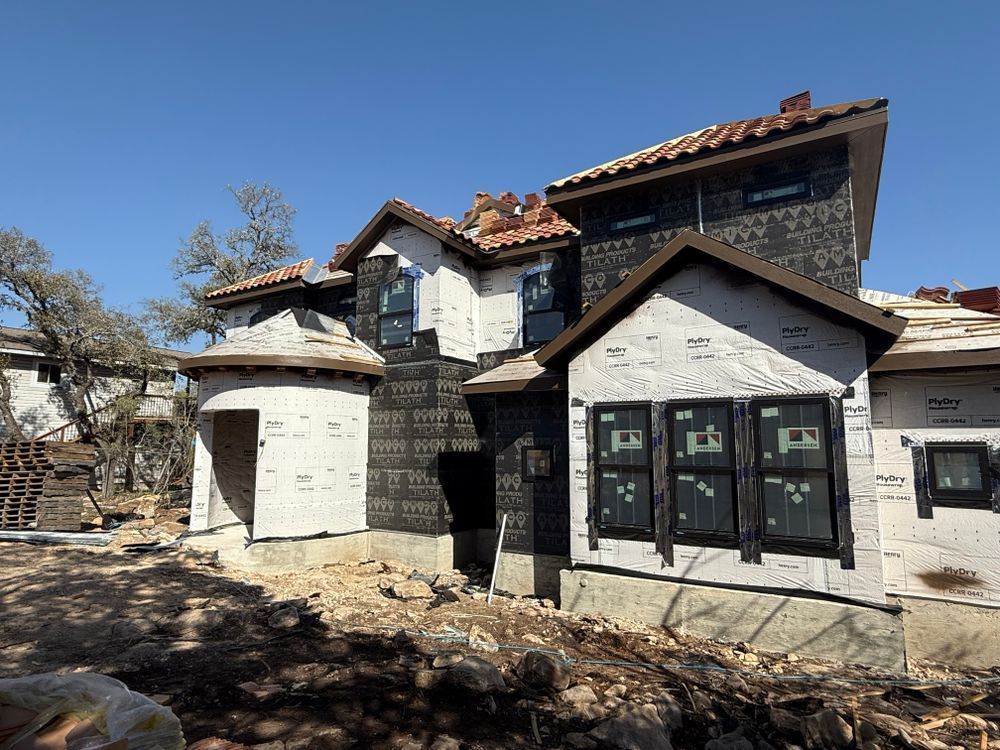 Stone and stucco house under construction with black-trimmed windows and red roof tiles on a sunny day