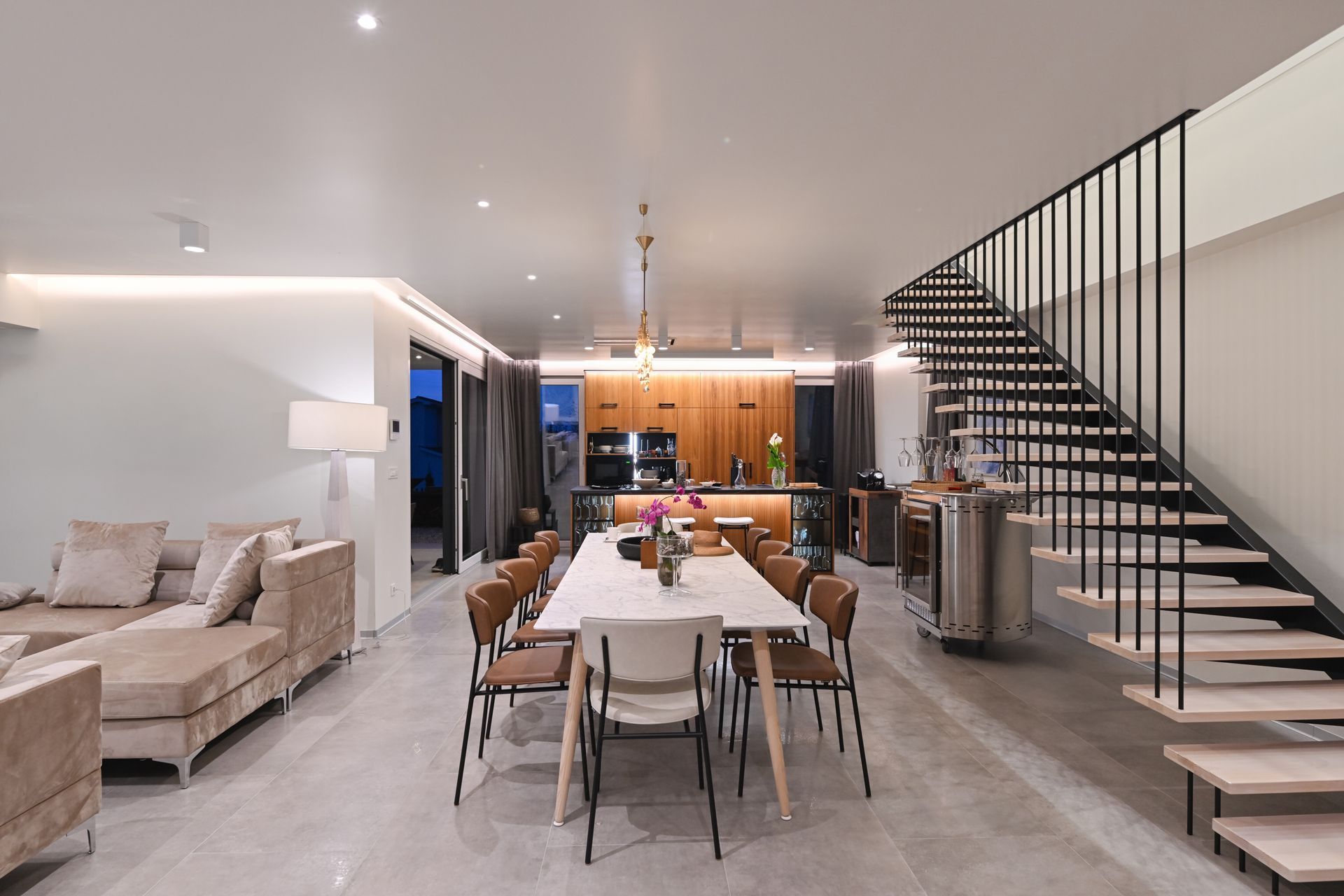 Modern open-plan living room with dining table, staircase, sofa, and warm kitchen lighting