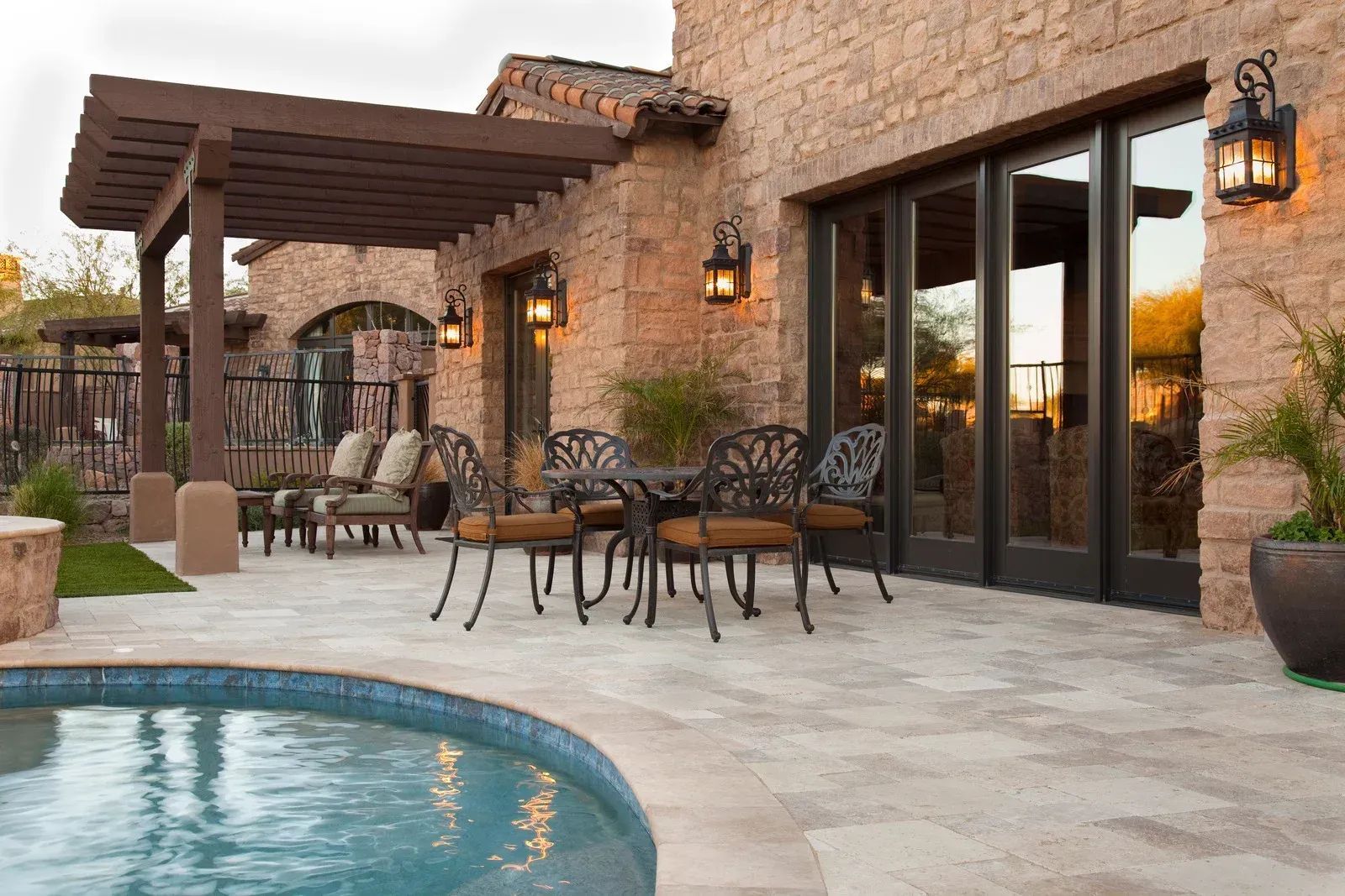 Patio with pool, stone building, pergola with seating, wrought iron table and chairs, and glass sliding doors.