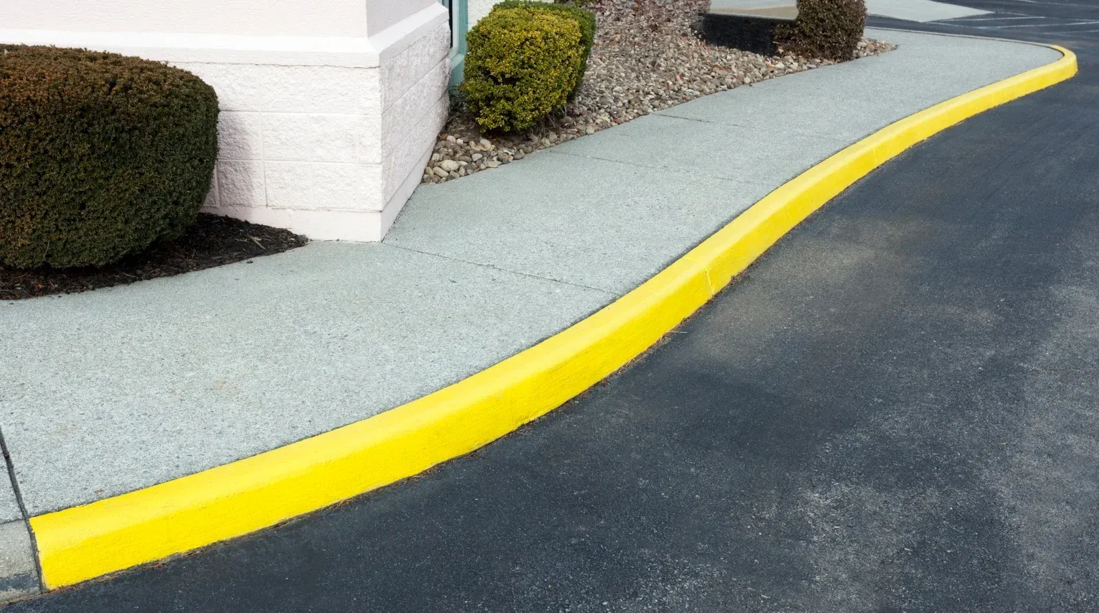 Yellow curb separates a gray walkway and a black asphalt driveway. Green bushes and white building are present.