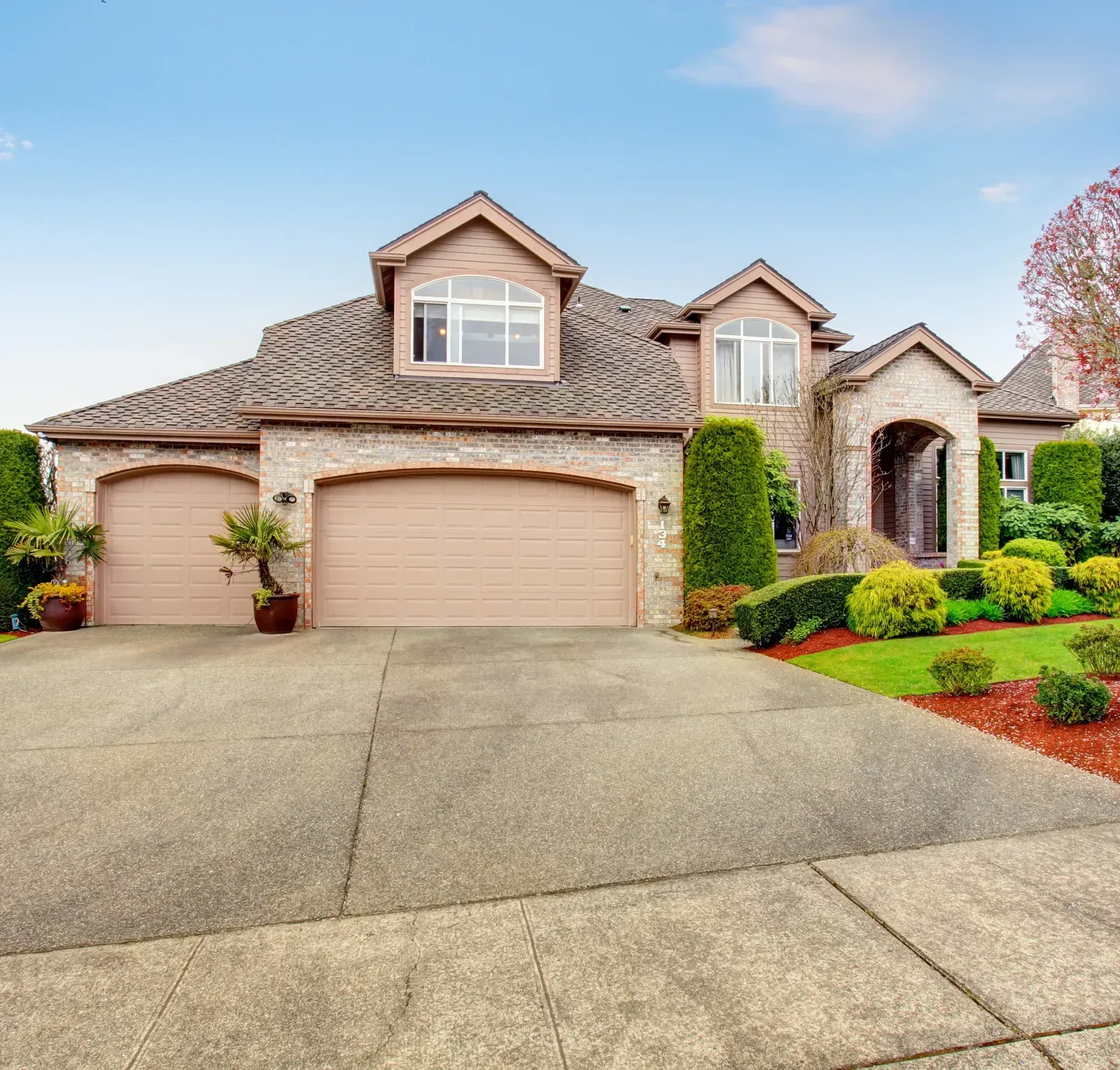 Tan brick house with a two-car garage and arched entrance, set on a concrete driveway and well-maintained lawn.