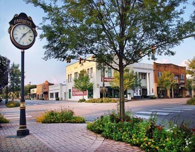 A street clock stands on a brick sidewalk in front of a tree-lined street with historic buildings in Fairhope, Alabama.