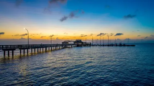 A long wooden pier stretches into calm blue water under a golden sunrise, with a structure at the end and distant masts.