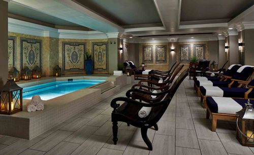A spa lounge with a rectangular pool, dark wood lounge chairs, mosaic wall art, and ambient lantern lighting.
