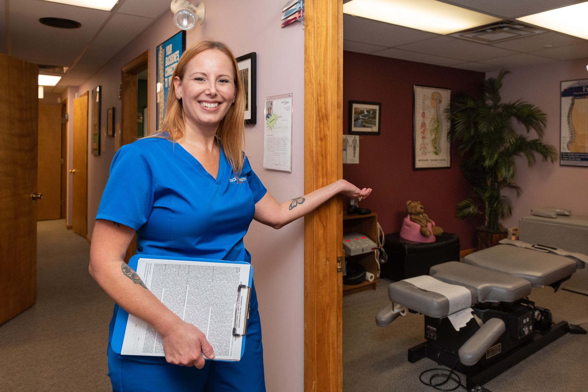 A woman in a blue scrub is standing in a hallway holding a clipboard.