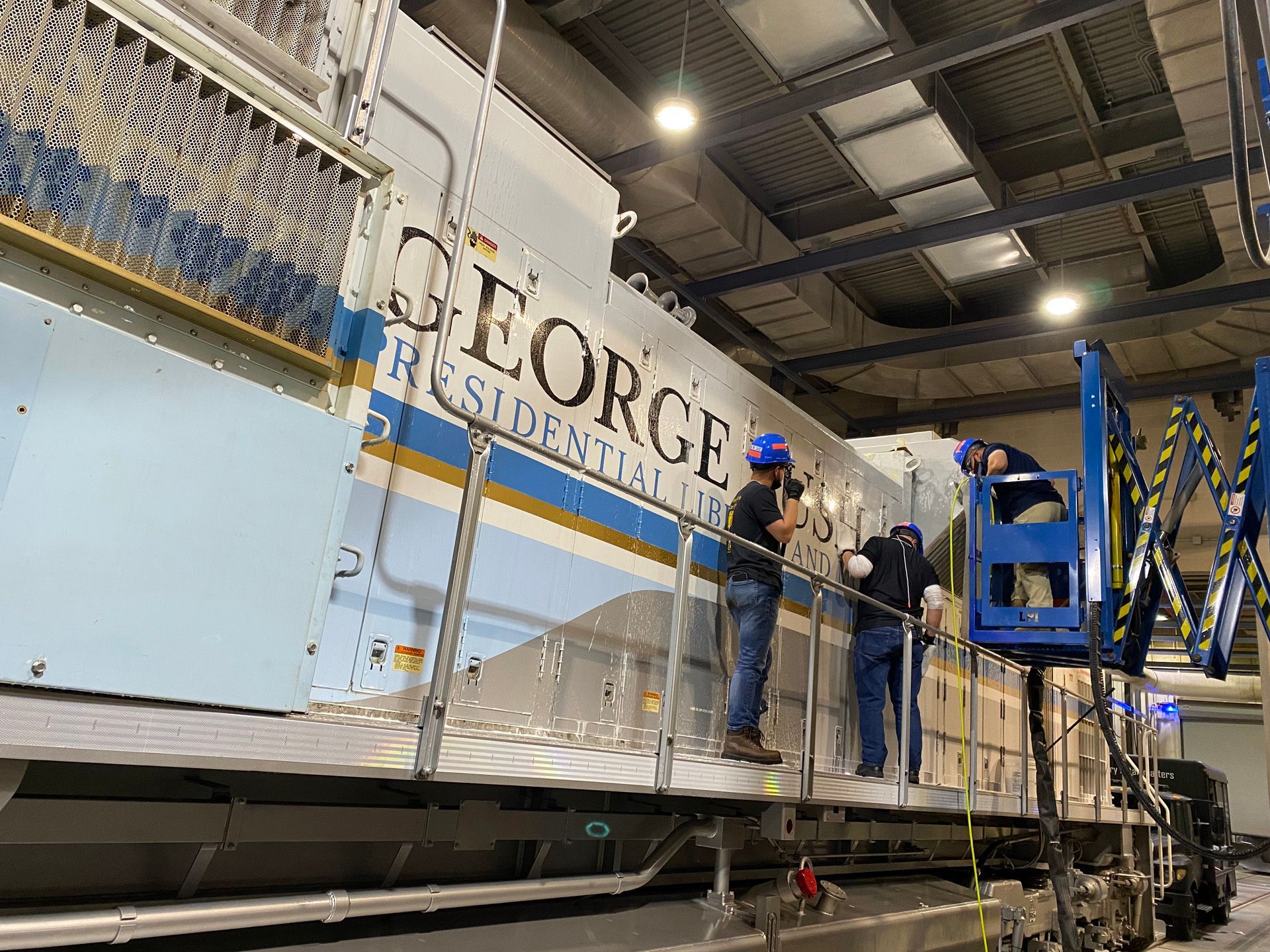 Workers on a light blue and white presidential train car, “GEORGE,” applying lettering. Indoors, industrial setting.