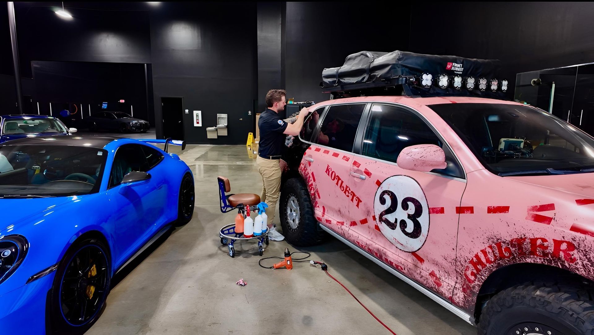 A man working on a pink Porsche SUV with a rooftop cargo carrier and a blue sports car in a shop.