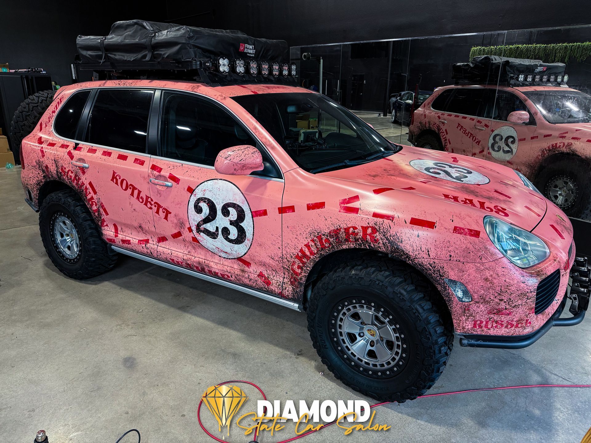 Pink off-road Porsche Cayenne with number 23 and roof rack at an event with another similar vehicle.