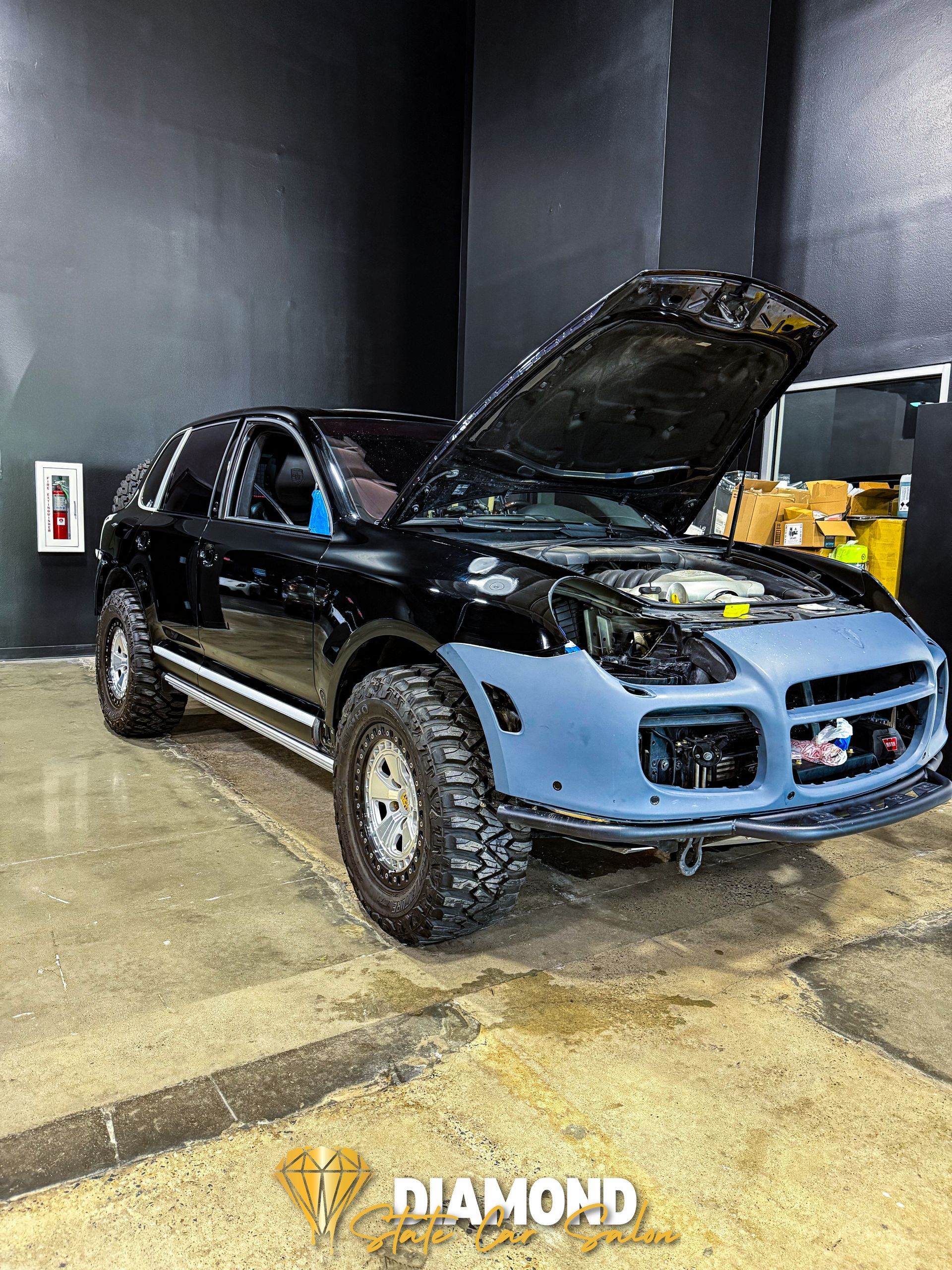 Black Porsche SUV with modified body, off-road tires, and hood open, in a garage.